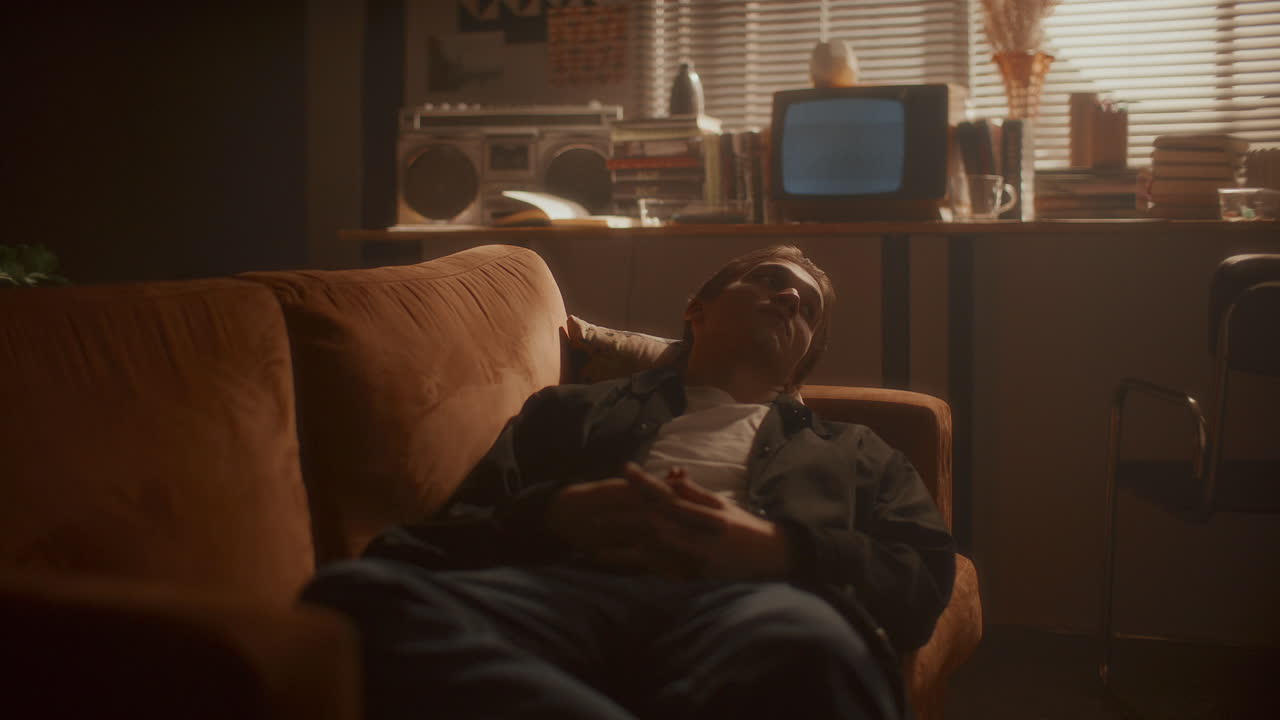 Young Pensive Man Resting on Sofa in Softly Lit Retro Room