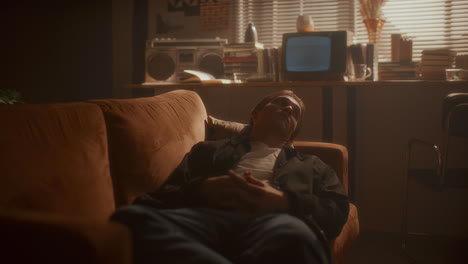 young pensive man resting on sofa in softly lit retro room