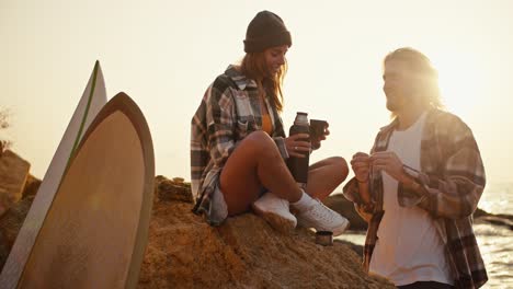 a blonde girl in a black hat and a plaid shirt sits on a large rock near the sea and opens a thermos of tea, and her blonde boyfriend looks at her. happy couple guy and girl relaxing on a rocky beach near the sea near their surfboards in the morning at sunrise