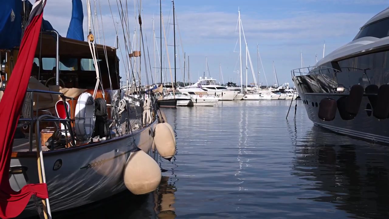 Vista panoramica del porto turistico con yacht di lusso, barche a vela e acque calme e riflettenti sotto un cielo azzurro brillante.