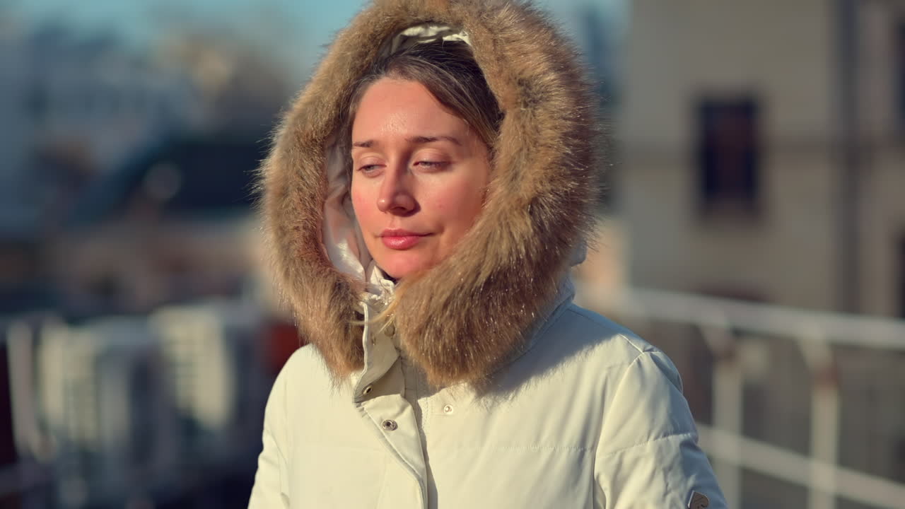 A woman relaxes on a rooftop, wearing a warm winter coat with a fur hood. She enjoys the bright sunshine and clear sky, creating a peaceful atmosphere