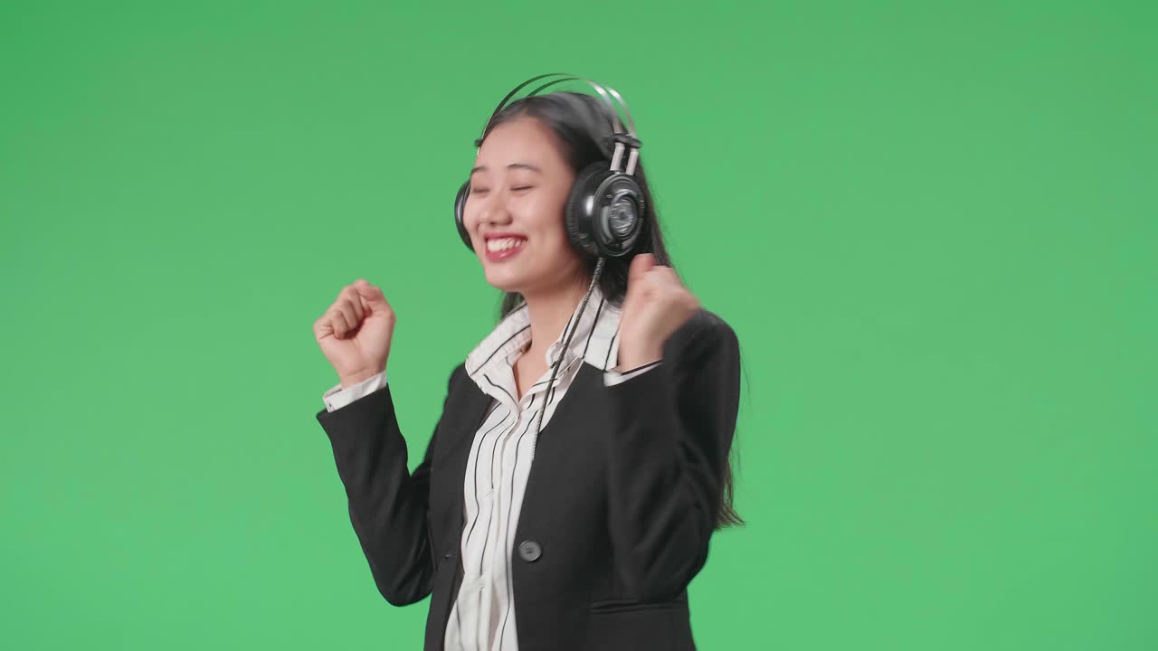 Side View Of An Asian Business Woman Listening To Music With Headphones And Dancing In The Green Screen Studio