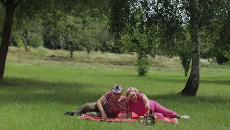 Family-weekend-picnic.-Active-senior-old-caucasian-couple-sit-down-on-blanket-on-green-grass-meadow