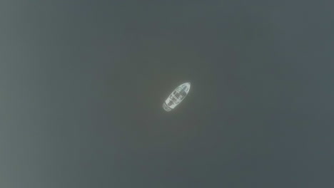 aerial ascends into thick fog cloud from lone boat on dark lake water