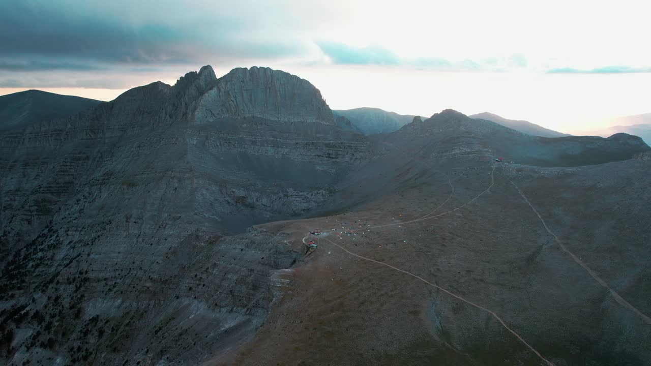 Ampia ripresa aerea di presentazione del monte Olimpo che rivela una capanna con campeggiatori e tende nella valle mentre il sole tramonta e le nuvole lo coprono.