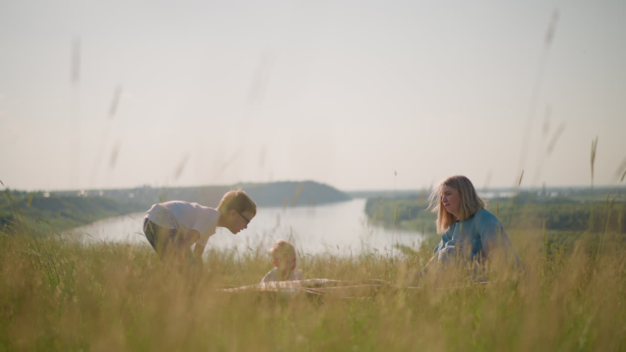 A woman in a blue gown and her sons in white shirts hold a large plaid scarf on a breezy hilltop beside a scenic lake. The family enjoys the fresh air and the natural beauty around them
