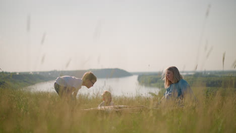 a woman in a blue gown and her sons in white shirts hold a large plaid scarf on a breezy hilltop beside a scenic lake. the family enjoys the fresh air and the natural beauty around them