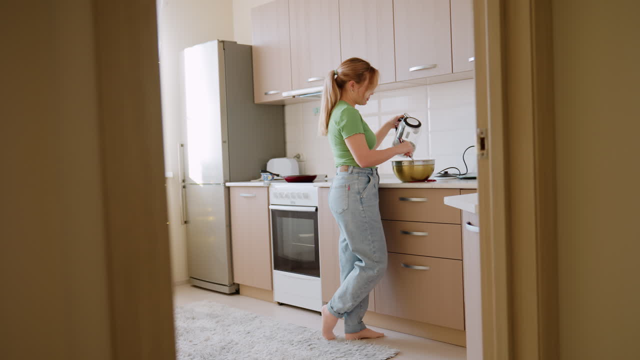 Housewife in green shirt and jeans pours water into mixing bowl while stirring ingredients during baking preparation, viewed through kitchen door with bright interior