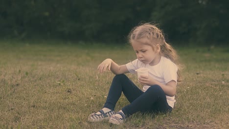 una niña con el pelo claro sostiene una taza de plástico y come yogur