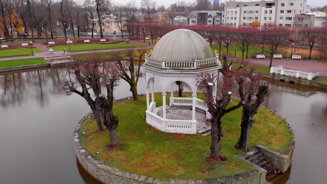 Riprese aeree ascendenti con drone che rivelano un pittoresco gazebo reale bianco situato al centro di un tranquillo laghetto all'interno di un parco pubblico