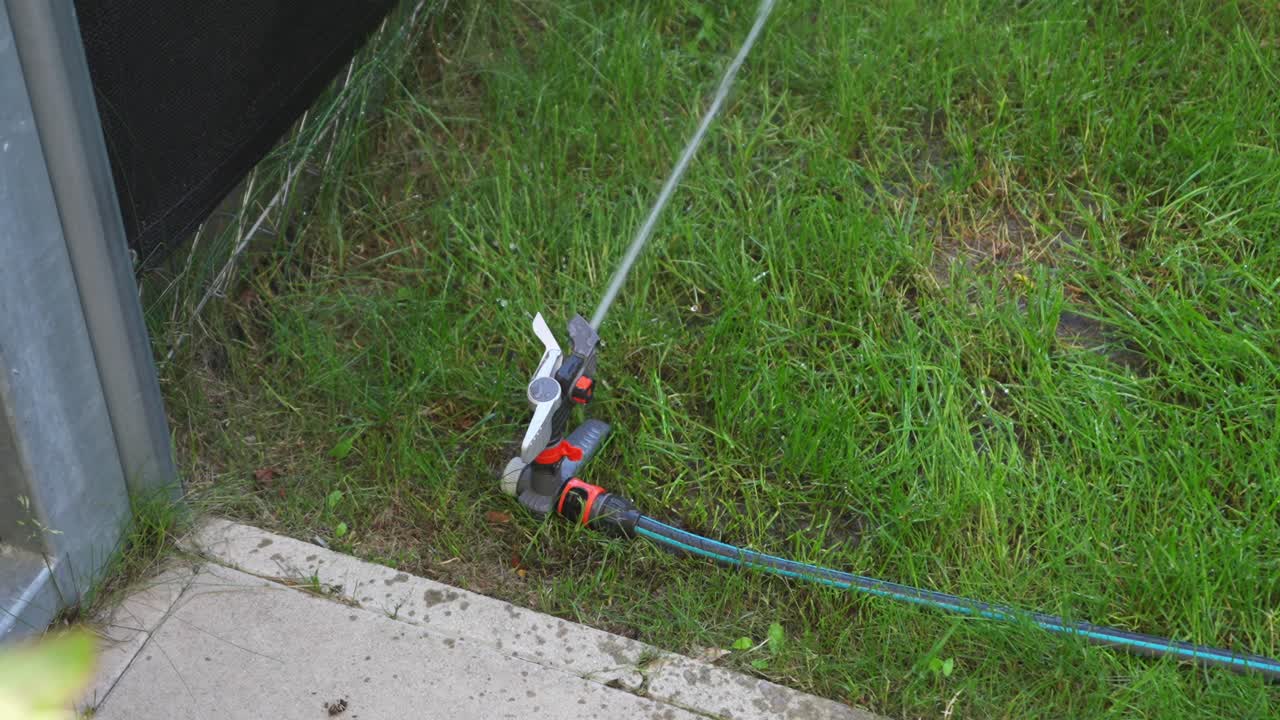 Orange gray impulse sprinkler in a lawn corner with green garden tube waters the lawn, view from diagonally high above