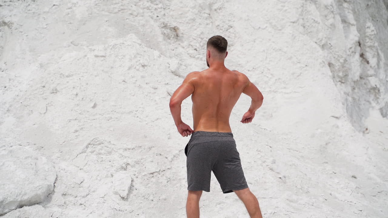 Backside view of a bodybuilder outdoors. Strong athlete without shirt shows his muscular body on white mountain background. Slow motion.