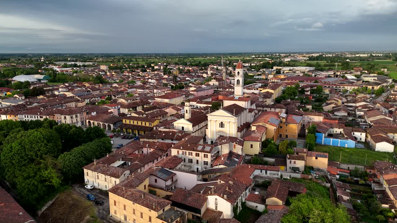 erial drone orbit of Pontevico, Lombardy, highlighting the Parish Church of Santa Maria Assunta with its tall bell tower, historic rooftops, narrow streets, and surrounding countryside at sunset
