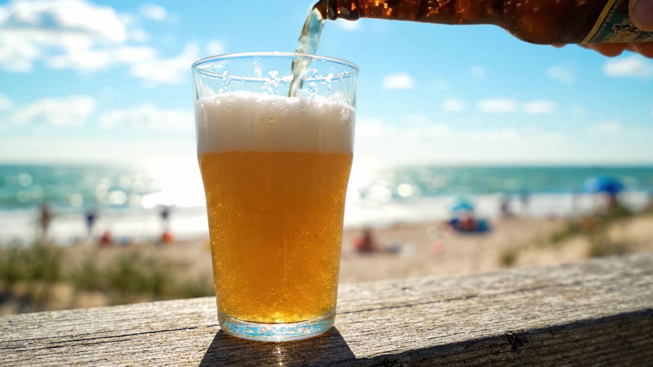 Momento Estivo Rinfrescante: Una Birra Fredda Versata in un Bicchiere su una Spiaggia Soleggiata, Catturando l'Essenza del Relax e del Piacere Tra le Onde e il Sole