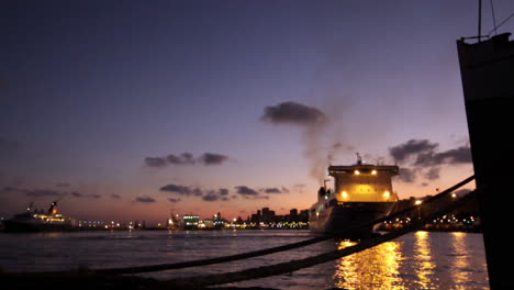 cargo ship illuminated at twilight, smoke rising as it departs the harbor