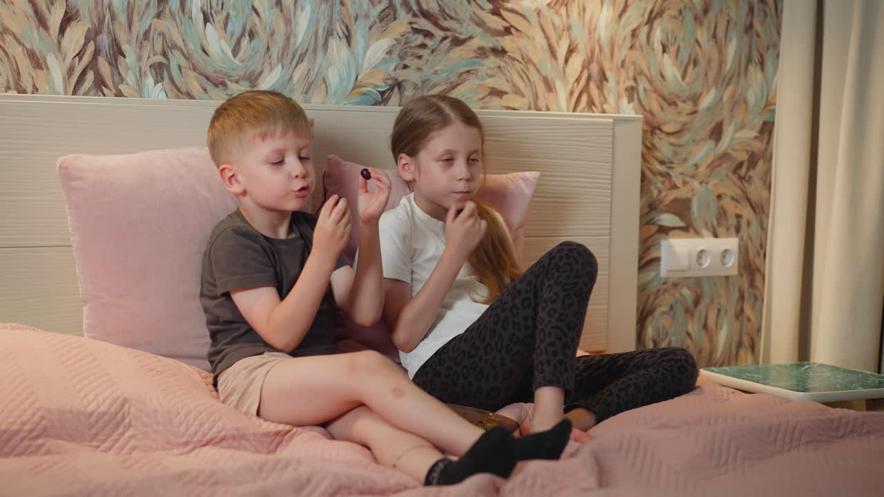brother and sister relaxing on bed eating black currant boy talking with sister cheerful moment cozy bedroom setting soft bedding colorful wallpaper electric outlet visible