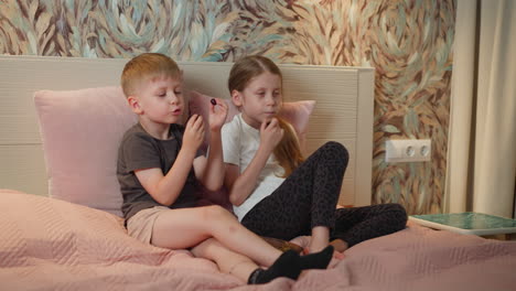 brother and sister relaxing on bed eating black currant boy talking with sister cheerful moment cozy bedroom setting soft bedding colorful wallpaper electric outlet visible