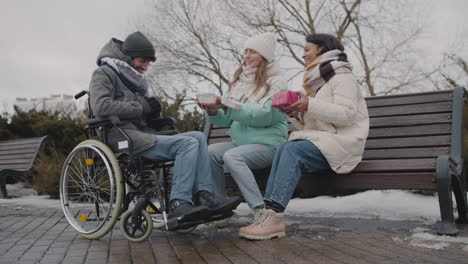 dos mujeres con un pastel pequeño y un regalo cantando feliz cumpleaños a su amigo discapacitado en el parque urbano en invierno