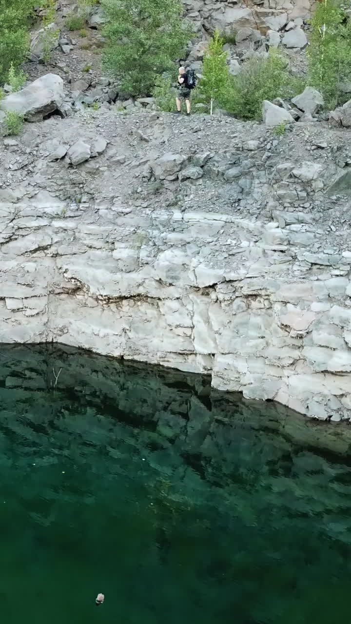 Aerial view of a young traveler with a backpack walking along a rocky path. Rocky shoreline of the lake with forest. Camping season Vertical video