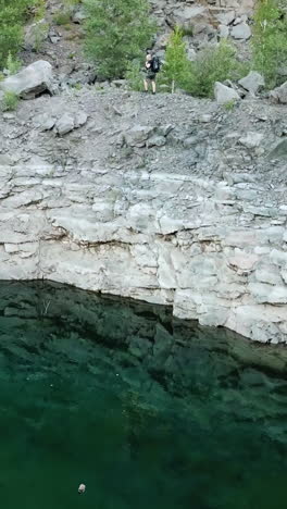aerial view of a young traveler with a backpack walking along a rocky path. rocky shoreline of the lake with forest. camping season vertical video