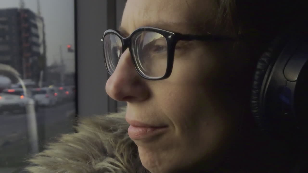Un'inquadratura statica cattura una persona con occhiali e cuffie, seduta su un tram in movimento a Budapest, con la scena della città riflessa nel finestrino.
