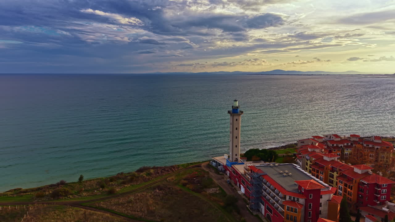 Il faro si erge maestoso accanto al mare calmo al tramonto, vicino a una vivace cittadina costiera con edifici colorati e un paesaggio pittoresco