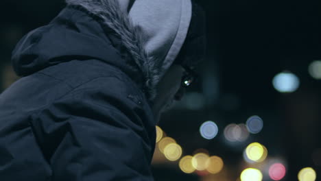 hooded man with glasses looks out over the city on a cold night