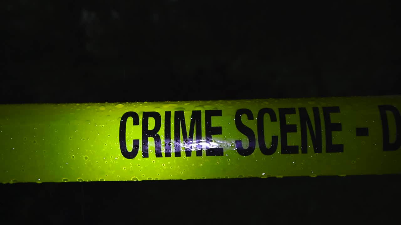 Primer plano de una cinta policial amarilla de «Escena del Crimen» colgada frente a una zona forestal oscura y totalmente negra al aire libre, mientras la lluvia y las gotas caen sobre la cinta a cámara lenta. Bien iluminado