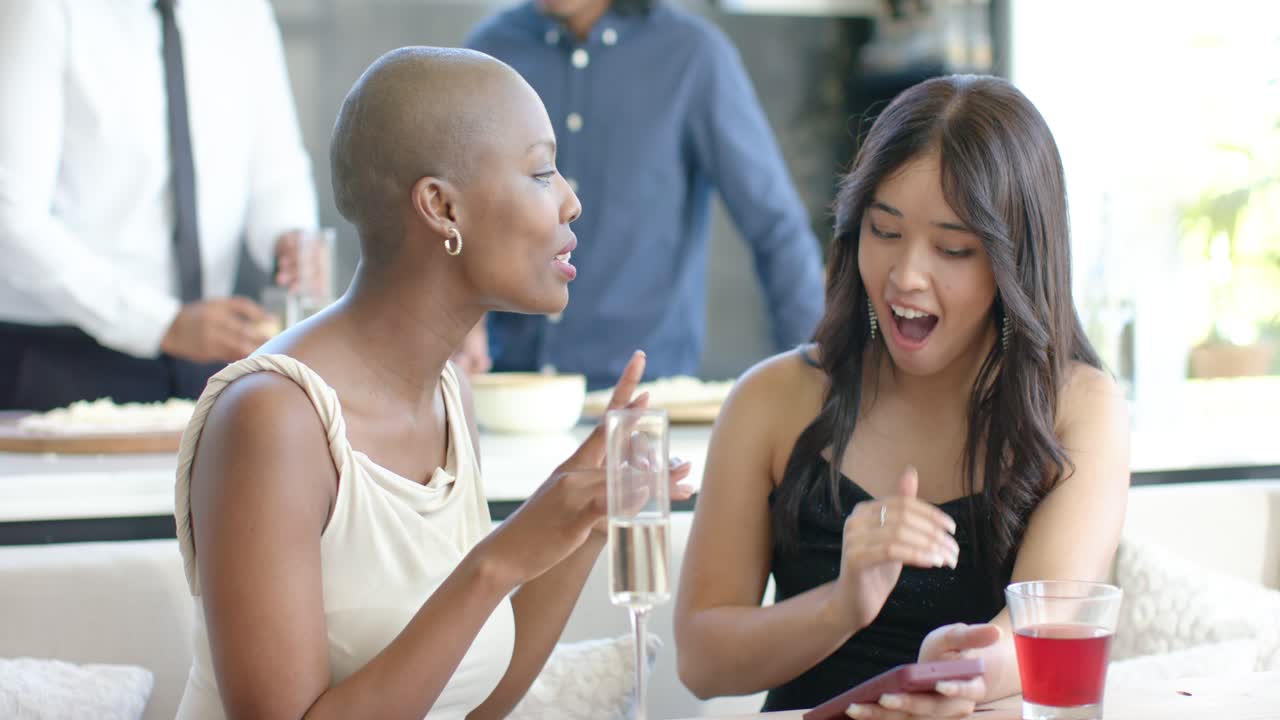 Two female friends leaning in as right friend placing smartphone showing funny post at wooden table