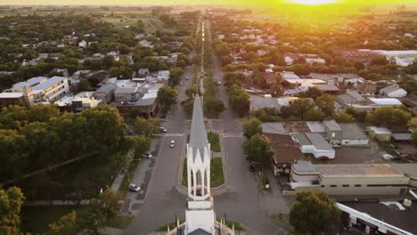 Aerial-view-flying-over-white-catholic-church-and-town-avenue-just-before-sunset