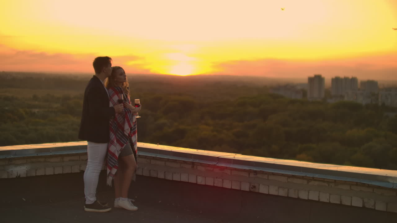 Giovane coppia sposata sul tetto che si abbraccia e beve vino rosso dai bicchieri, in piedi e vestita a quadri, ammirando il bellissimo tramonto sulla città.