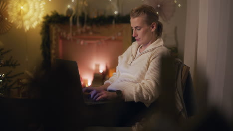 a woman works on a laptop at night writing an email against the background of christmas tree decorations and lights