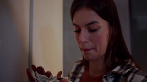 Close-up-a-happy-brunette-girl-in-a-checkered-shirt-takes-out-a-piece-of-pie-in-foil-from-the-refrigerator-and-greedily-eats-it-with-pleasure-at-night-in-a-modern-apartment