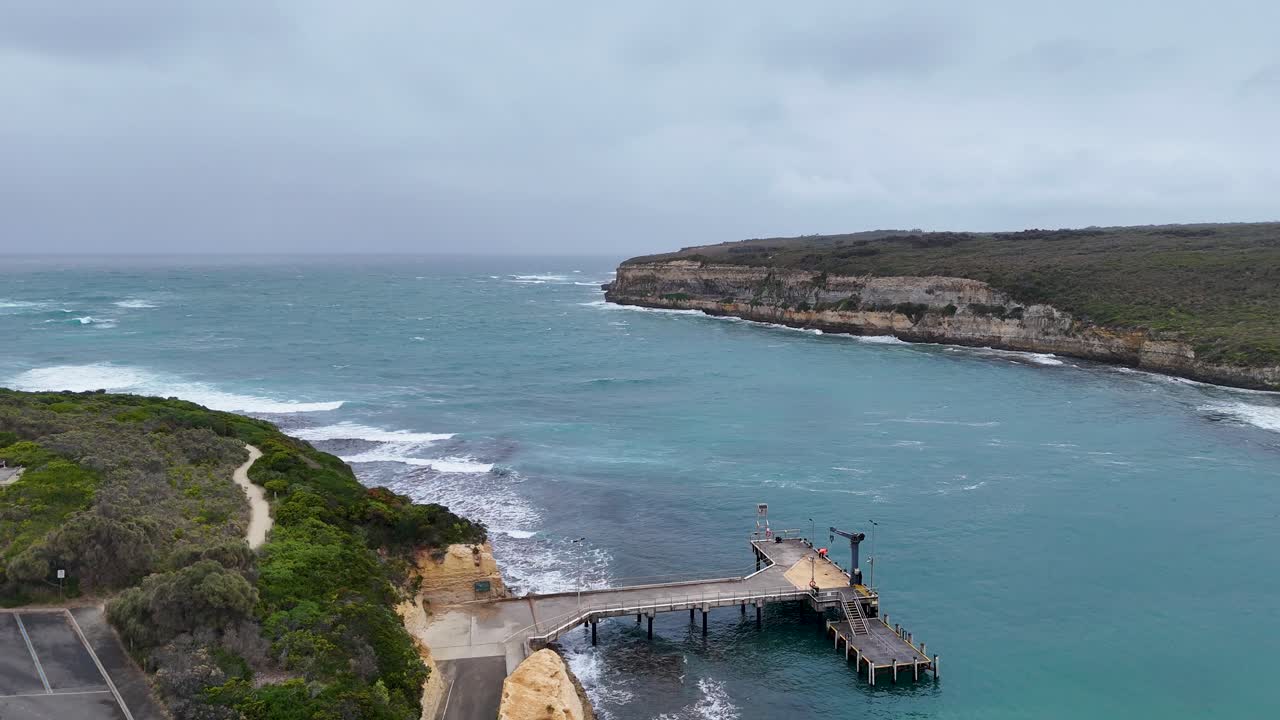 Drone footage captures Port Campbell's rugged coastline and pier under overcast skies, highlighting ocean waves and natural erosion