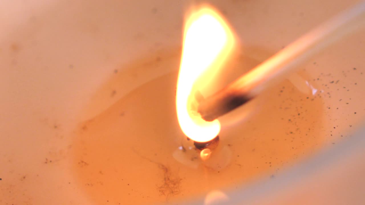Filmato macro al rallentatore di uno stoppino di candela che viene acceso da un fiammifero di legno per bruciare con una fiamma arancione brillante che tremola nella cera di soia.