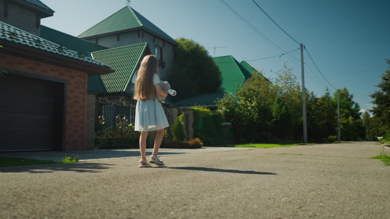 Rear view of little girl in white dress with long flowing hair holding teddy bear while walking down empty residential street on sunny day with tree shadows cast on pavement and houses on both sides