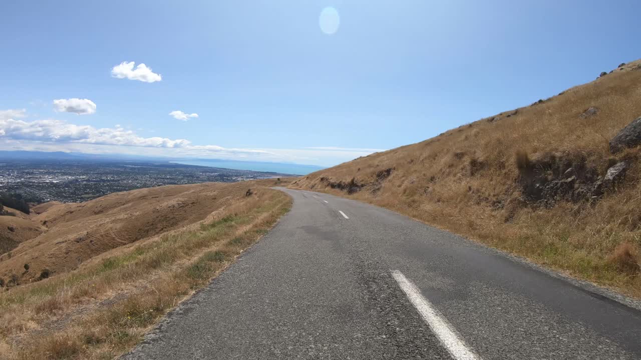 POV from car bonnet driving on hill road with city and clouds in background.