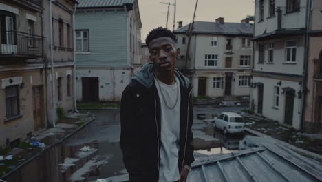 young confident african american male standing on rooftop, wearing hooded jacket and chain, overlooking decaying urban courtyard with gritty architectural backdrop at dusk