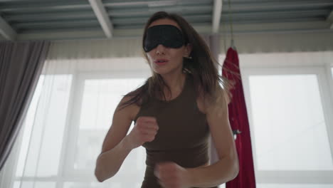 energetic trainer blindfolded her eyes and punches air with intensity and focus during solo session inside modern yoga studio, practicing controlled motion and strength with heightened body awareness