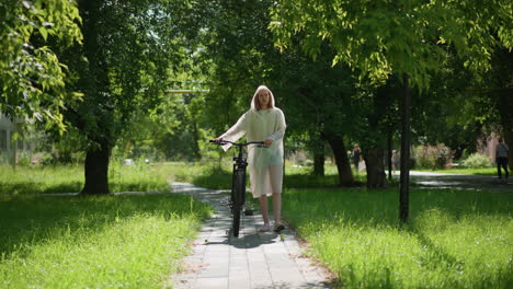 young woman in translucent raincoat strolls along sunlit pathway with her bicycle, pausing to place stand and remove hood, background features vibrant greenery, trees, and distant pedestrians