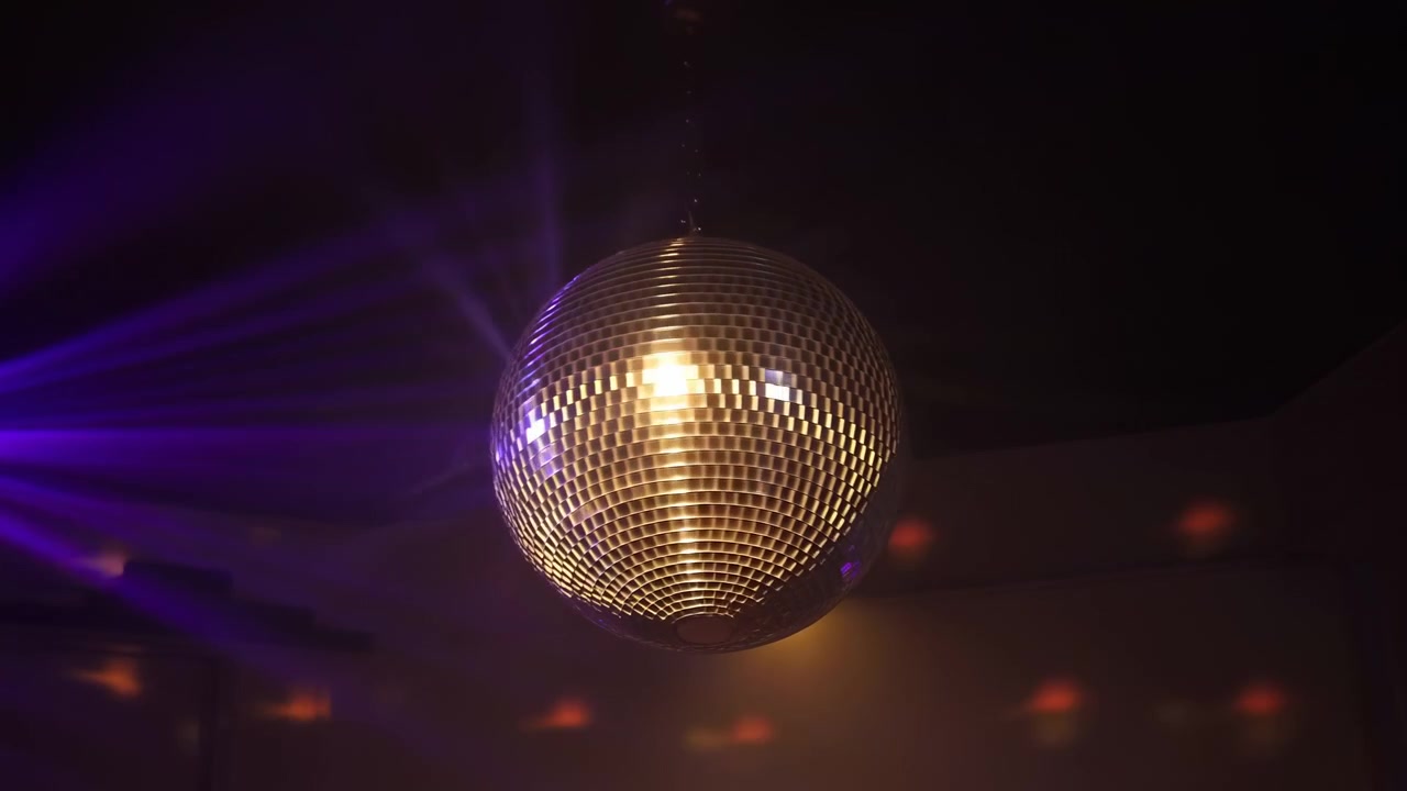 Una palla da discoteca a specchi pende dal soffitto in una discoteca buia, riflettendo raggi di luce viola e calda attraverso l'interno e creando un'atmosfera di festa.