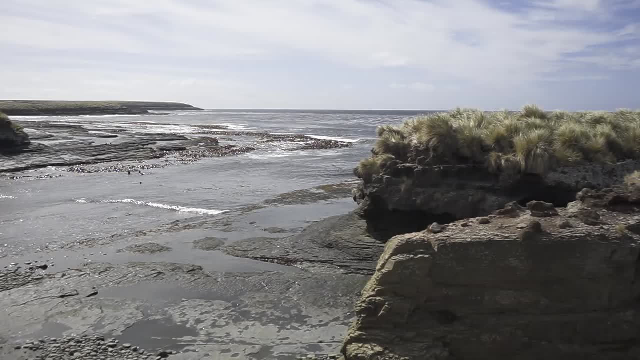 Ocean waves crashing against a rocky coastline with sparse vegetation, gradually zooming in on a prominent rock formation