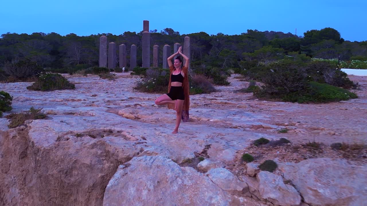 Woman performing a yoga tree pose on a rocky cliff during a serene sunset in Cala Llentia Ibiza. Majestic aerial view flight fly push forward drone