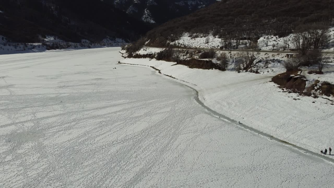 Drone flying slowly over frozen Pineview Reservoir in Northern Utah in wintertime, with people walking their dog on shoreline