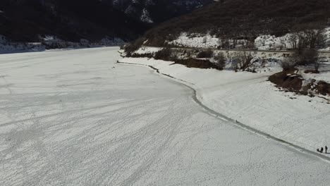 Drone-flying-slowly-over-frozen-Pineview-Reservoir-in-Northern-Utah-in-wintertime,-with-people-walking-their-dog-on-shoreline