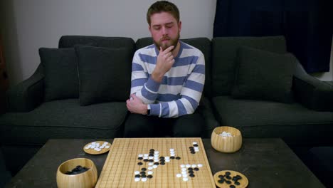 beaded man strokes beard while thinking of strategic move in go board game on bamboo board on stone table by grey couch in living room