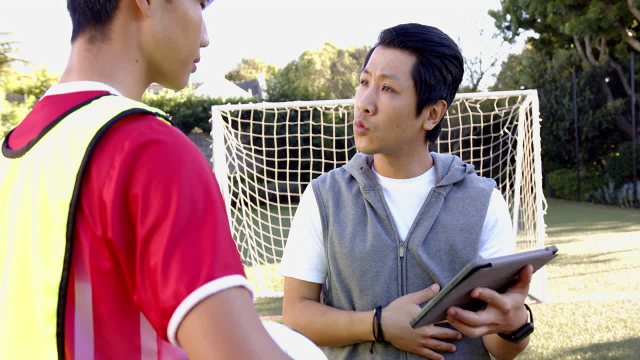 Asian father holding tablet talking to his teenage son in a soccer field