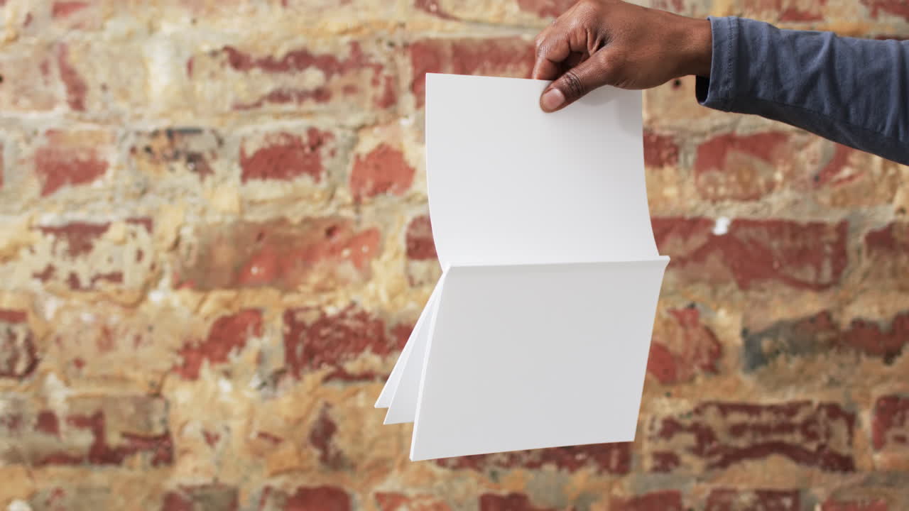 Video of hand of african american man holding book with blank pages, copy space over brick wall