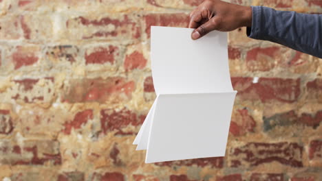 Video-De-La-Mano-De-Un-Hombre-Afroamericano-Sosteniendo-Un-Libro-Con-Páginas-En-Blanco,-Copiando-Espacio-Sobre-Una-Pared-De-Ladrillo