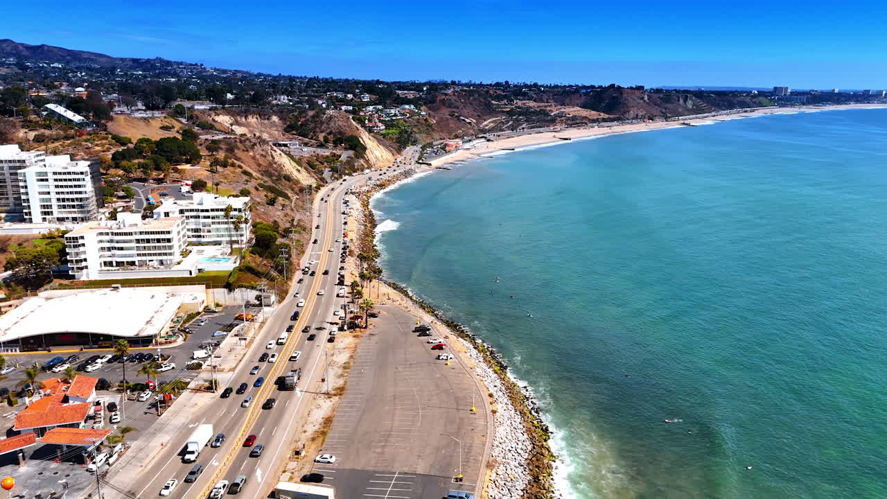 Numerous cars move by the highway along the narrow sandy beach. Drone footage over the coast of the Pacific Ocean in Malibu, California, USA